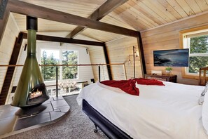 A bedroom with a bed, a fireplace, a TV, and a window with a view of trees. - Big Bear Gambrel #2066 (Big Bear Lake)
