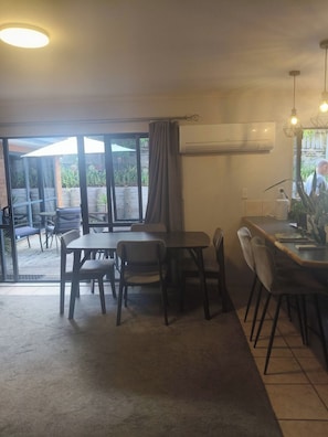A dining area with a table and chairs, an air conditioning unit on the wall, and a view through large windows.