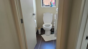 Ein schmaler Flur, der zu einem Badezimmer mit Toilette, Fenster und Mülleimer führt.