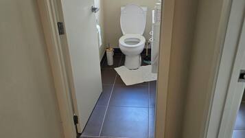 A narrow hallway leading to a bathroom with a toilet, a window, and a trash bin.