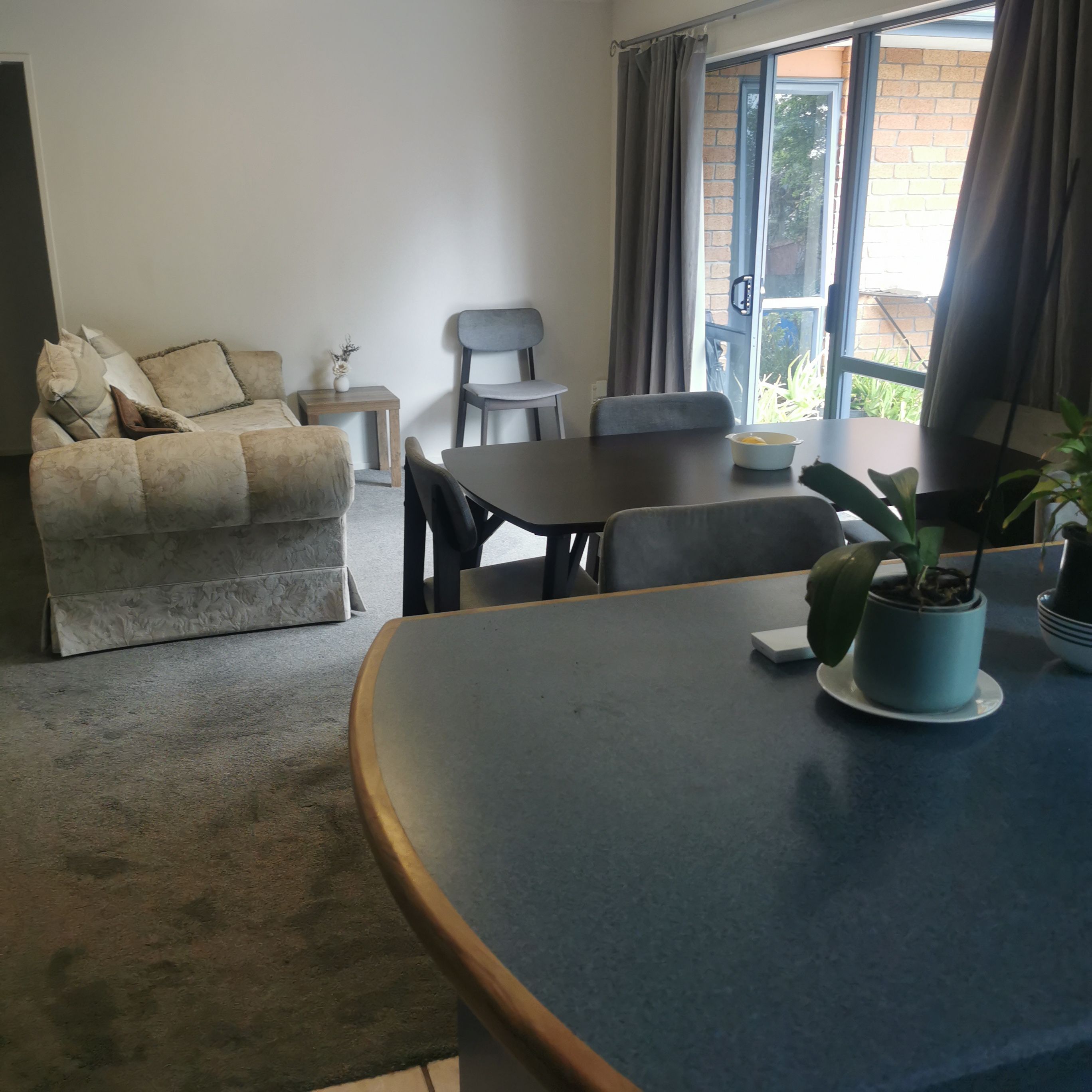 A living room with a sofa, a dining table, chairs, a small table with a vase, and a window with curtains.