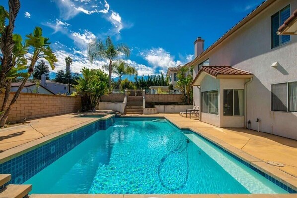 A swimming pool with a clear blue water, surrounded by a patio area with steps leading to a house.