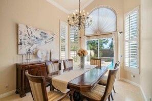 A dining room with a long table, chairs, a chandelier, a vase with flowers, and a large window with shutters.