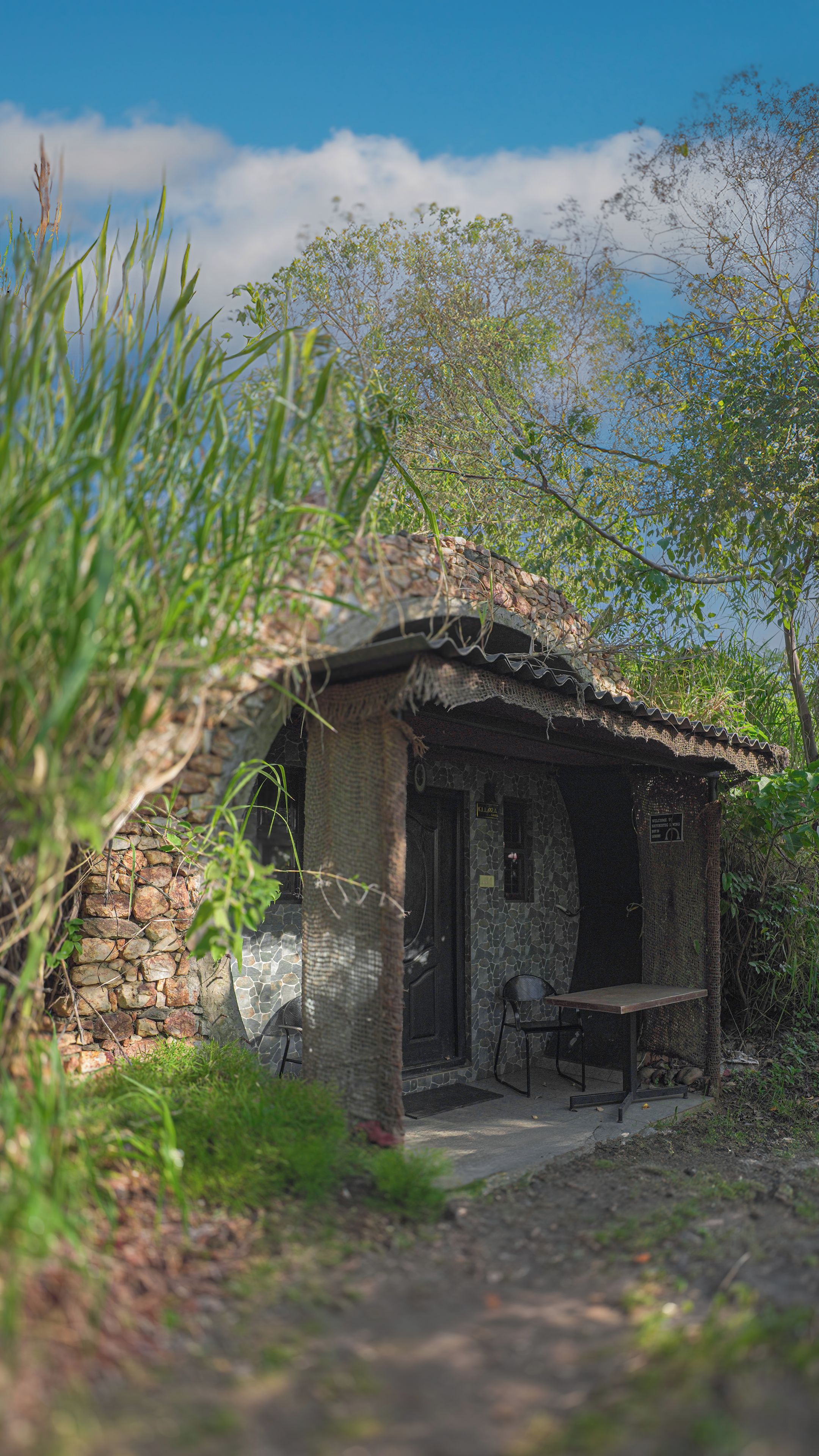 Un edificio rústico de piedra con techo de tejas, un banco de madera afuera y una pared de piedra parcialmente cubierta por follaje.