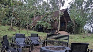 A rustic outdoor seating area with metal chairs and a fire pit, surrounded by greenery and a small building with a sloped roof.
