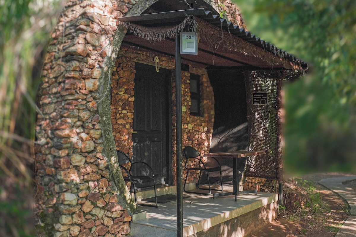 Una estructura de piedra rústica con puerta de madera, un banco y una mesa, rodeada de follaje.
