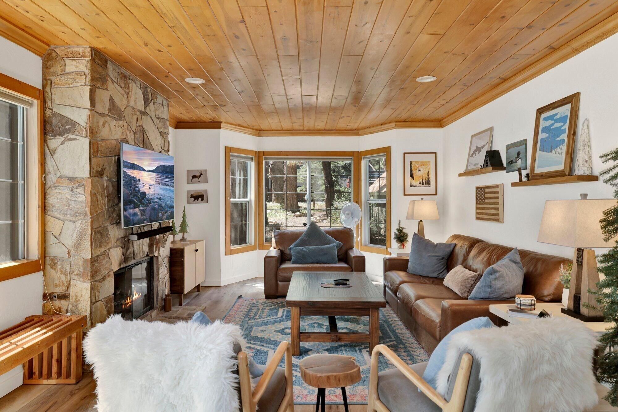 Un salon chaleureux avec une cheminée en pierre, un plafond en bois et de grandes fenêtres donnant sur l’extérieur.