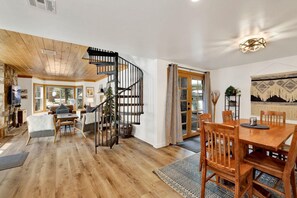 A spacious living area with a wooden floor, a fireplace, a dining table with chairs, and a spiral staircase. - Green Canoe Cabin #2293 (Big Bear Lake)