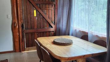 A wooden dining table with chairs, a woven placemat, and a view of a forest through a window with curtains.