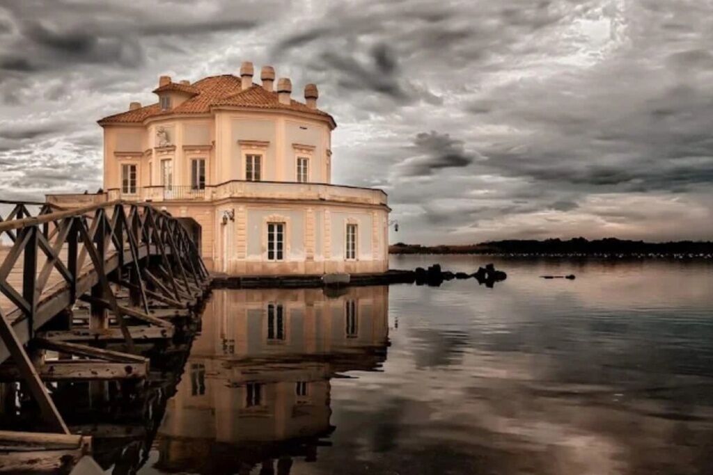 A historic building with a water tower, situated by a calm body of water under a cloudy sky.