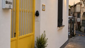 Een smalle straat met een wit gebouw, voorzien van een geel hekwerk, een zwart luik en een kleine potplant.
