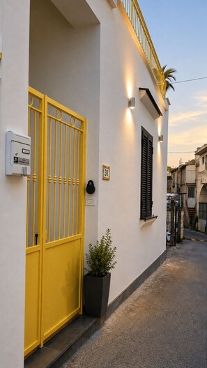 Eine schmale Straße mit einem weißen Gebäude, das ein gelbes Tor, ein Fenster mit schwarzen Rollläden und einen kleinen Blumentopf zeigt.