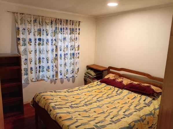 A bedroom with a bed, a wooden headboard, a nightstand, a shelf, and a window with floral curtains.