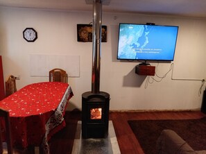 A living room with a TV mounted on the wall, a wooden table with a red tablecloth, a clock, and a wood-burning stove.