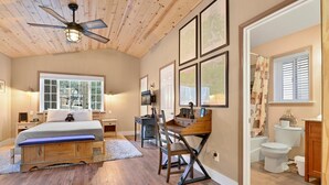 Un dormitorio con cama, escritorio y silla, ventilador de techo y ventana con vista a árboles.