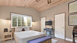 Un dormitorio con una cama grande, un banco de madera, un escritorio con una computadora, un televisor y obras de arte enmarcadas en las paredes.
