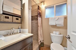 A bathroom with a shower, a toilet, a sink with toiletries, and a window with towels. - Whispering Woods #2375 (Big Bear Lake)