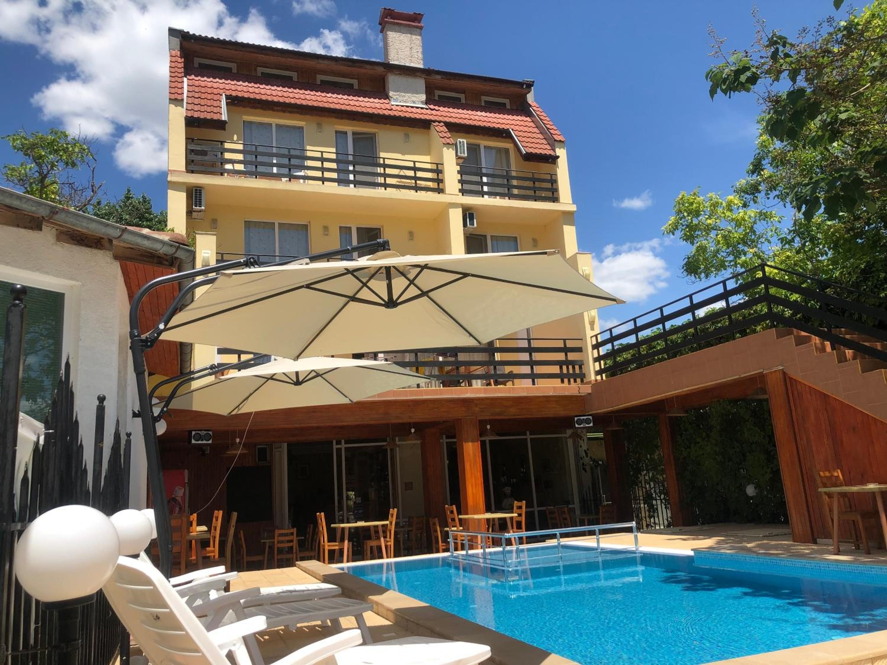 A swimming pool with lounge chairs and umbrellas, a building with balconies, and a clear blue sky.