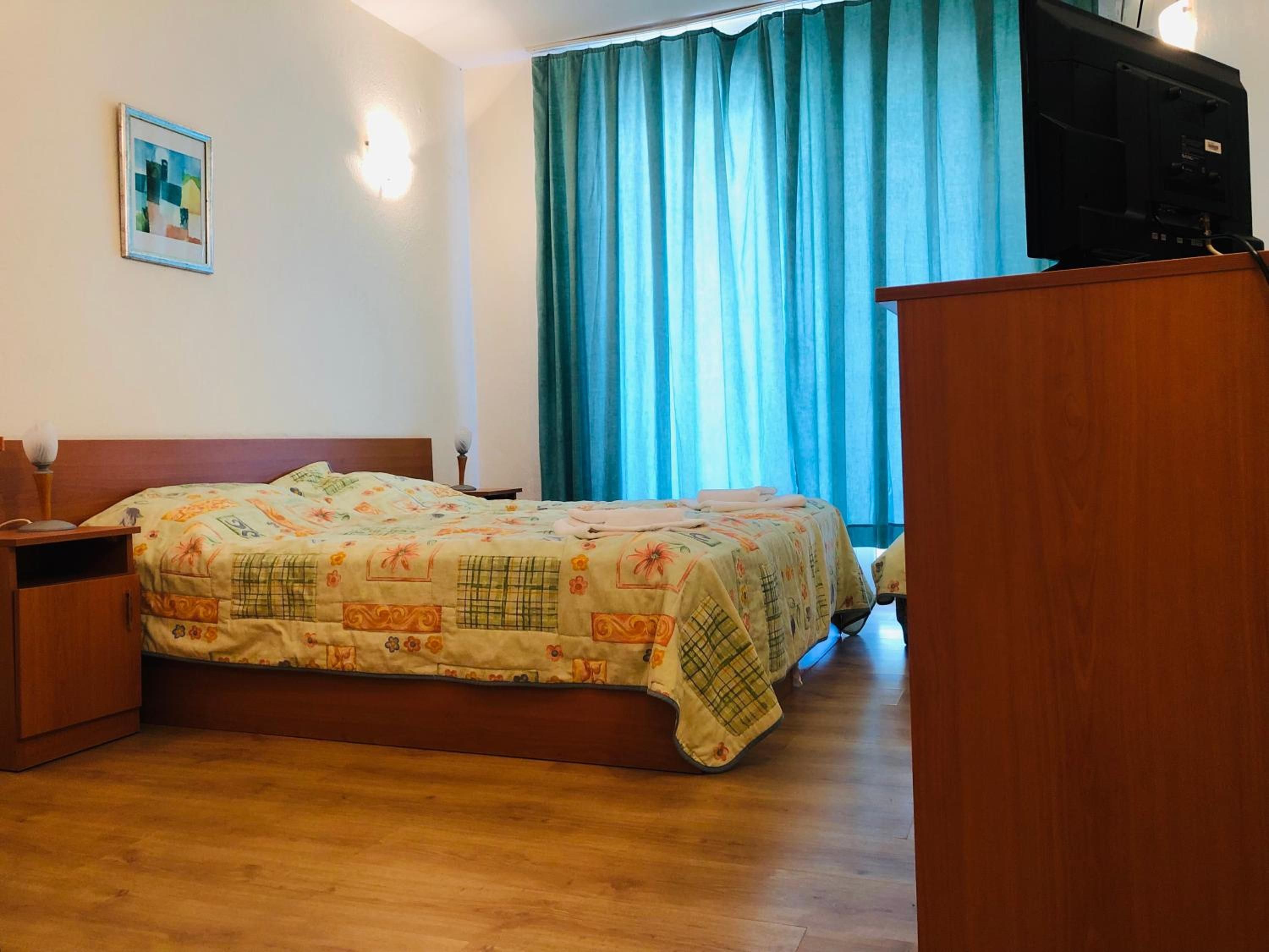 A hotel room with a bed, a wooden nightstand, a desk, a television, and a window with curtains.