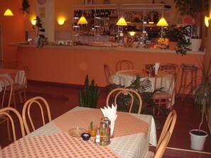 Interno di un ristorante con zona bar, tavoli e sedie, e piante in vaso.