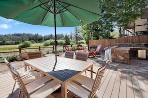 An outdoor patio with a wooden table and chairs, a green umbrella, and a stone fireplace. - Big Bear Den #2152 (Big Bear Lake)