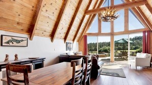 A spacious room with a high wooden ceiling, a dining area with a wooden table and chairs, and a view of a green landscape through large windows.
