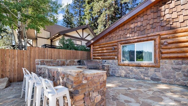 A log cabin with a stone patio, outdoor grill, and white plastic chairs.