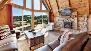 A spacious loft-style room with a stone fireplace, leather furniture, and large windows offering a view of a green landscape.