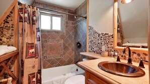A bathroom with a stone mosaic wall, a bathtub, a toilet, and a sink with a wooden countertop.
