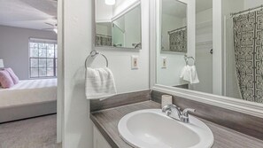 A bathroom with a large mirror, a sink, and a shower with a patterned curtain. There is a towel hanging on the wall and a bed visible in the adjacent room. - Landing Jacksonville (Jacksonville)