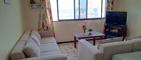 A living room with a sofa set, a coffee table with a vase, a television on a stand, and a window with a view of the sea.