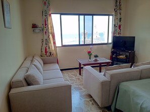Un salon comprenant un canapé, une table basse avec un vase, un téléviseur posé sur un meuble et une fenêtre donnant sur la mer.