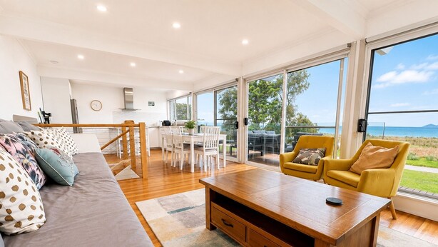A spacious living area with a sofa, coffee table, and yellow armchairs. A kitchen is visible in the background with a dining table and chairs. - Coastal 3-Bed Haven With Panoramic Views (Portarlington)