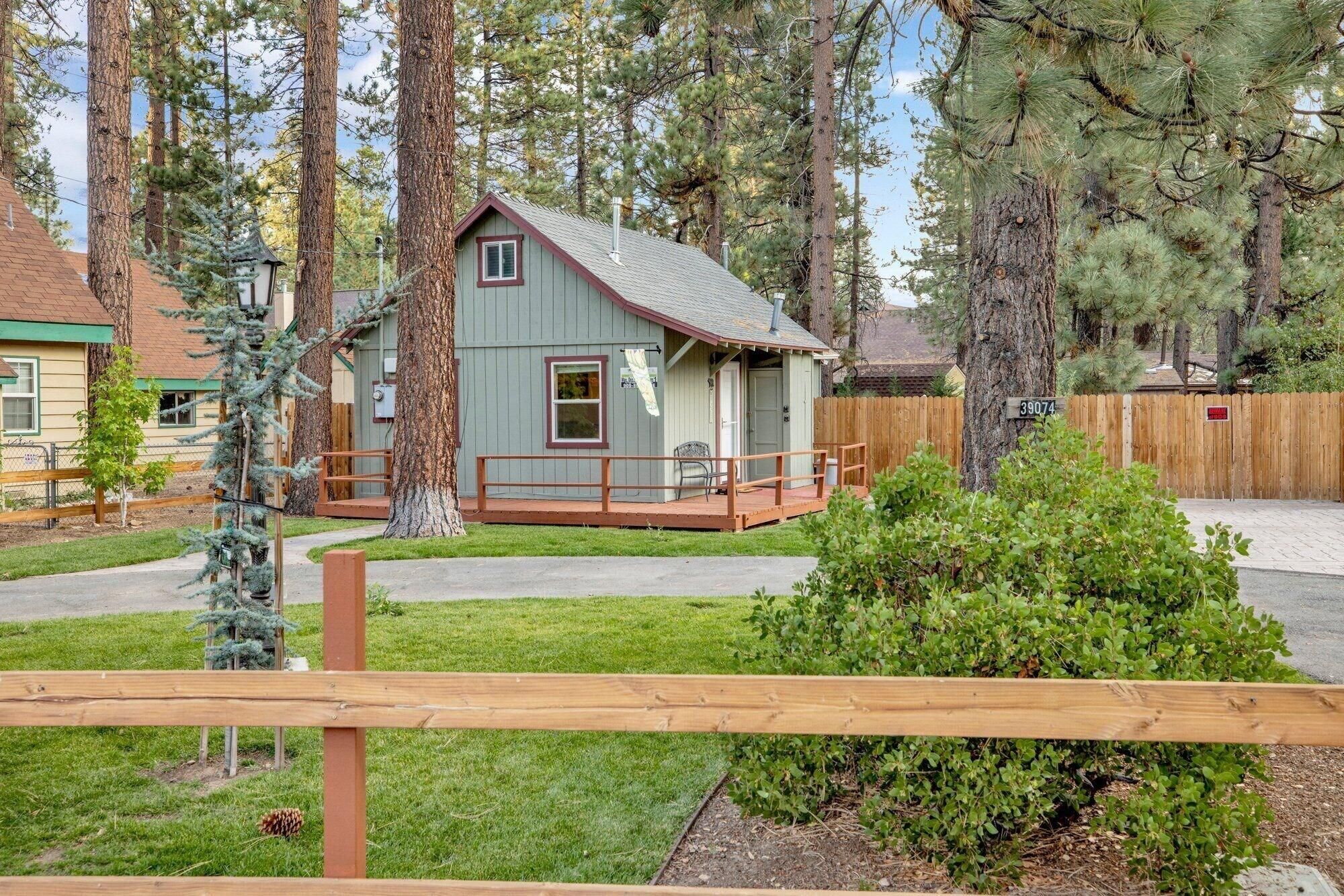 A small, single-story cabin with a porch, surrounded by tall trees and a wooden fence.