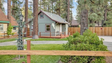 A small, single-story cabin with a porch, surrounded by tall trees and a wooden fence.