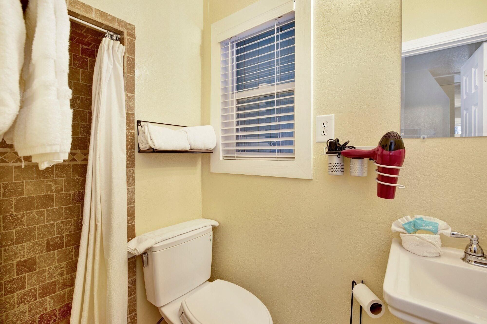 A bathroom with a toilet, a shower curtain, a towel rack, a window with blinds, and a hairdryer mounted on the wall.