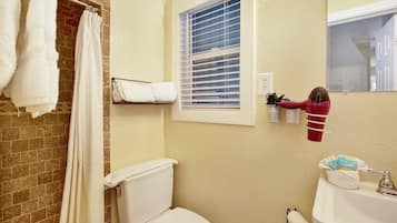 A bathroom with a toilet, a shower curtain, a towel rack, a window with blinds, and a hairdryer mounted on the wall.