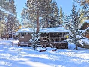 A cabin with a wooden deck and stairs leading to a porch, surrounded by snow-covered trees. - Sunny Bear Cabin #1970 (Big Bear Lake)