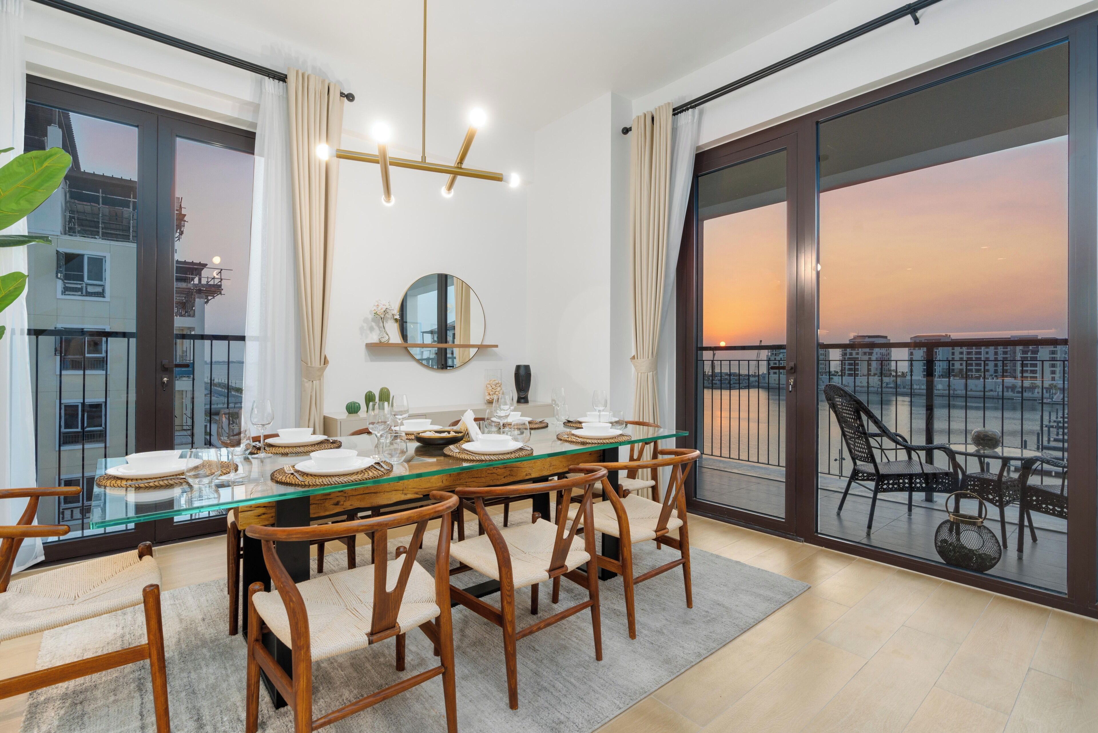 A dining area with a glass table, wooden chairs, and a view of the sunset through large windows.