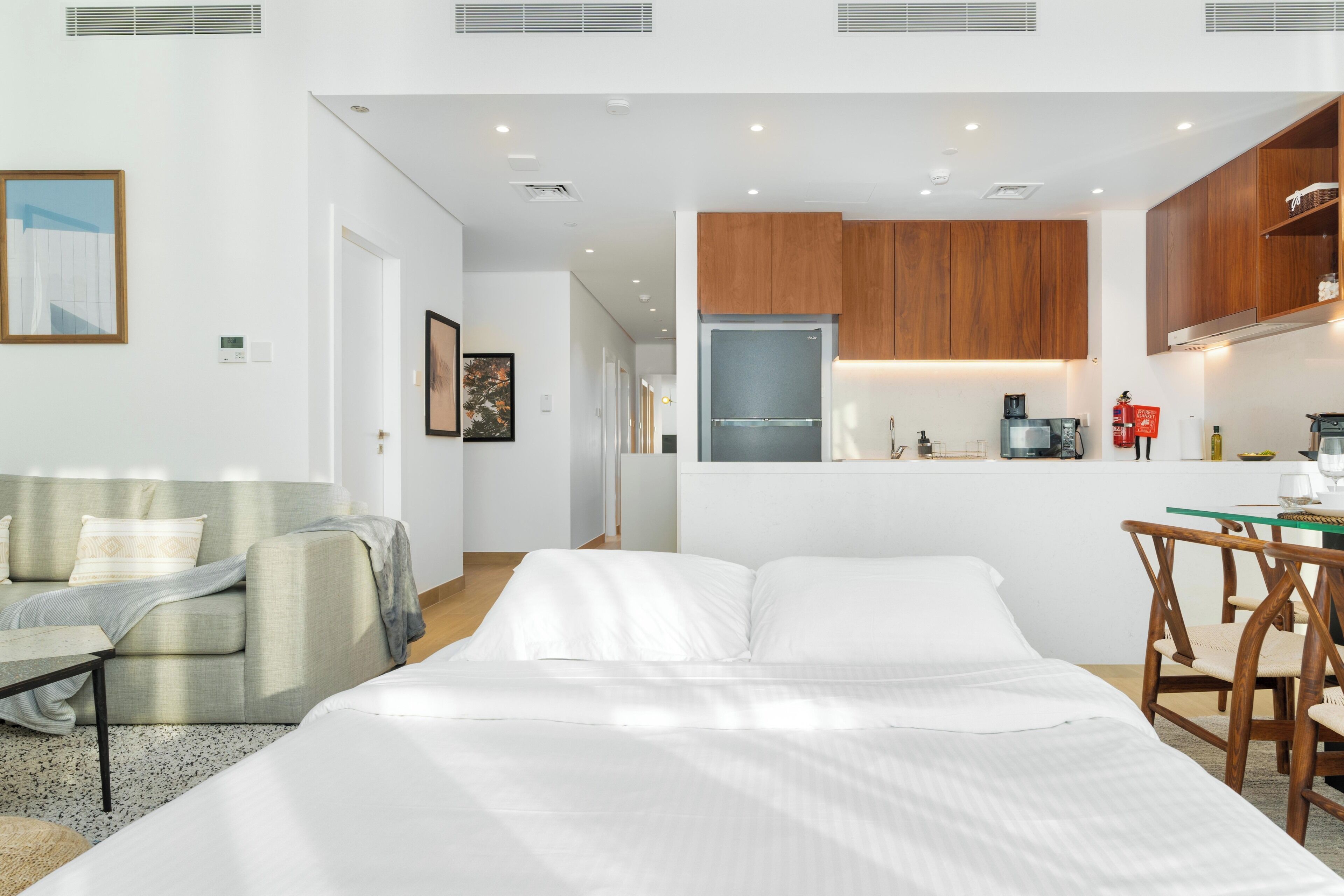 A modern hotel room with a bed, a seating area with a sofa and armchair, a kitchenette with wooden cabinets, and a dining table with chairs.