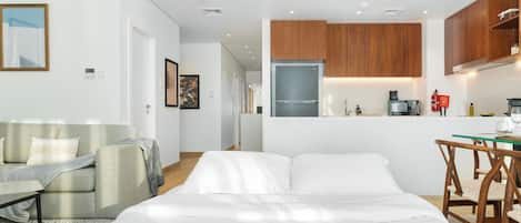 A modern hotel room with a bed, a seating area with a sofa and armchair, a kitchenette with wooden cabinets, and a dining table with chairs.