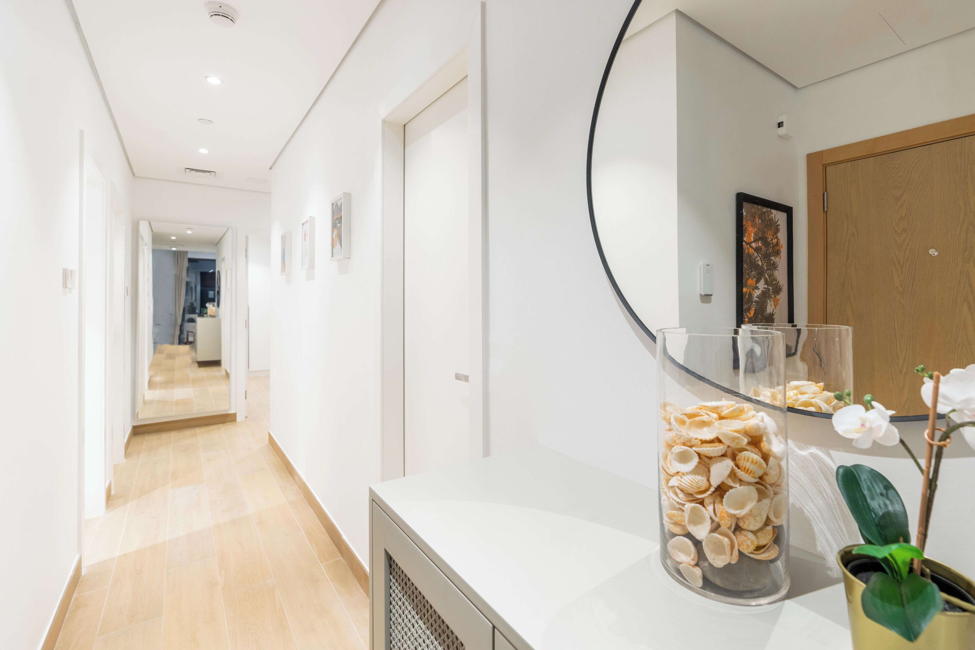 A modern hallway with a white wall, a wooden door, and a glass vase with seashells on a white counter.