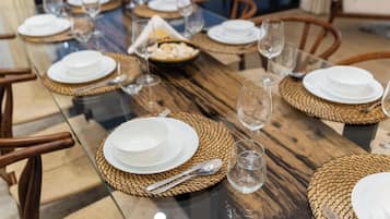 Una mesa de comedor dispuesta con platos blancos, cubiertos y vasos de vidrio transparente, rodeada de individuales tejidos y sillas de madera.