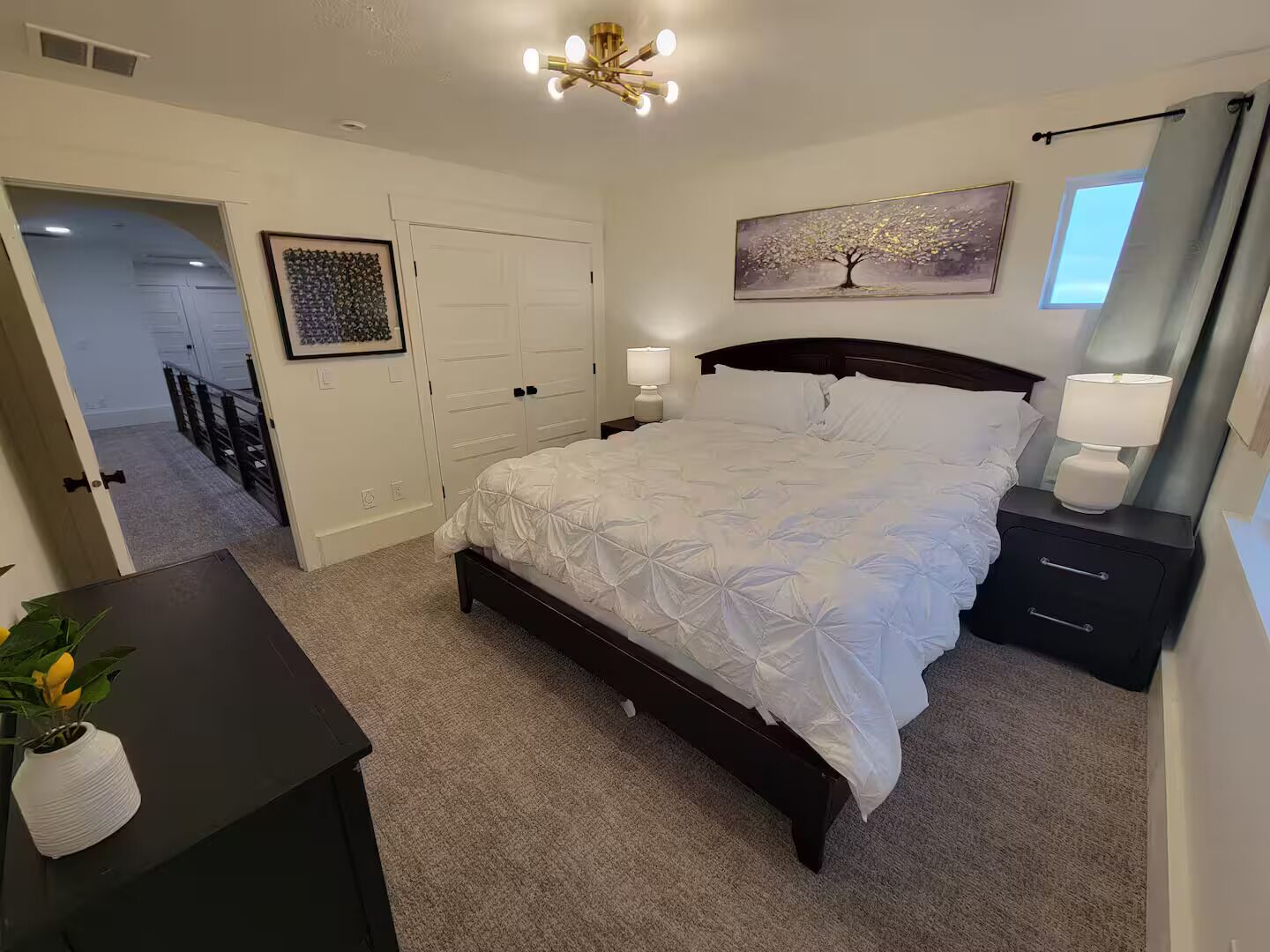 A bedroom with a bed, bedside tables, a nightstand, a ceiling light fixture, a door, and a wall-mounted painting.