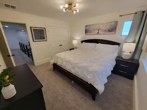 Un dormitorio con una cama, mesitas de noche, lámpara de techo, una puerta y un cuadro colgado en la pared.