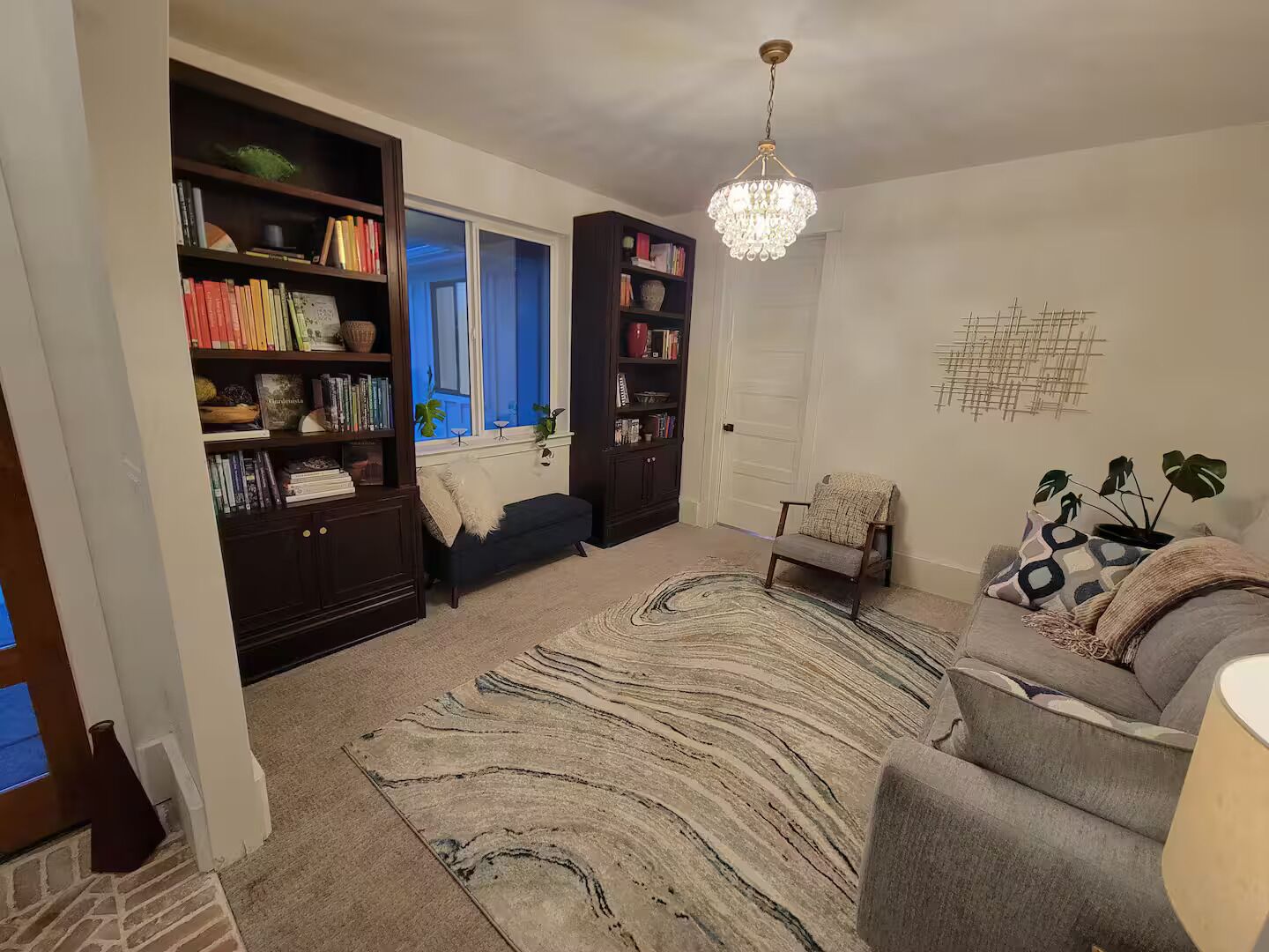 A living room with a bookshelf, a sofa, a chair, a rug, and a chandelier.