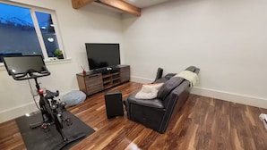 Sala de estar com piso de madeira, um sofá preto, um rack para TV e uma bicicleta ergométrica.
