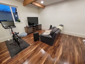 A living room with a wooden floor, a black sofa, a TV stand, and a stationary bike. - Beautiful, spacious country home in Cache Valley with views (Wellsville)
