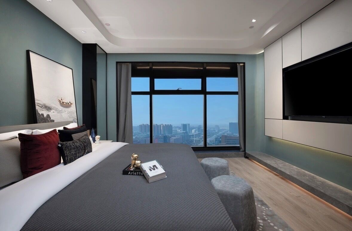 A modern bedroom with a large bed, a flat-screen TV mounted on the wall, and a city view through the window.