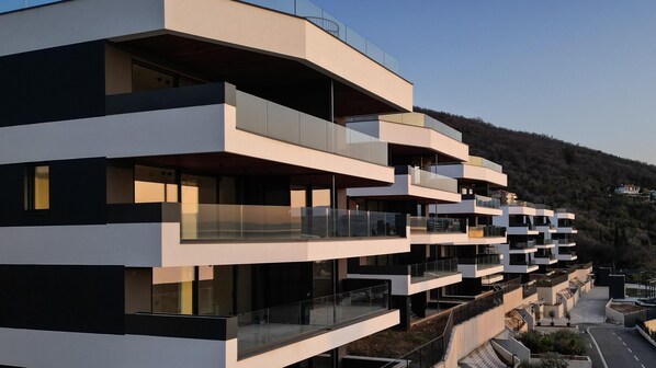 A multi-story residential building with balconies, a clear sky, and a hilly landscape in the background.